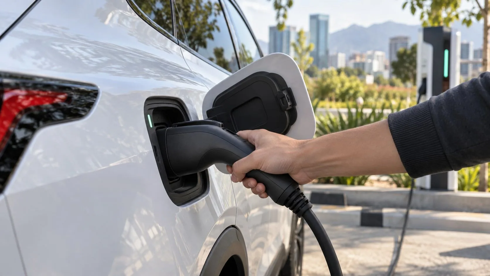 Mano sujetando un conector de carga conectado al puerto de un auto eléctrico blanco en una estación.